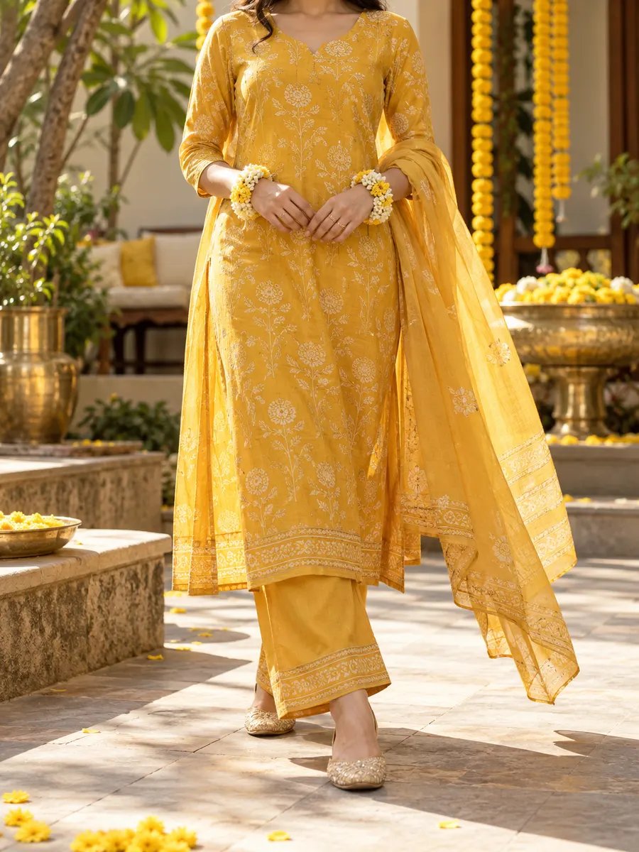 A woman in a marigold kurta, palazzos and a sheer yellow dupatta at a haldi, garlands of marigolds behind her.