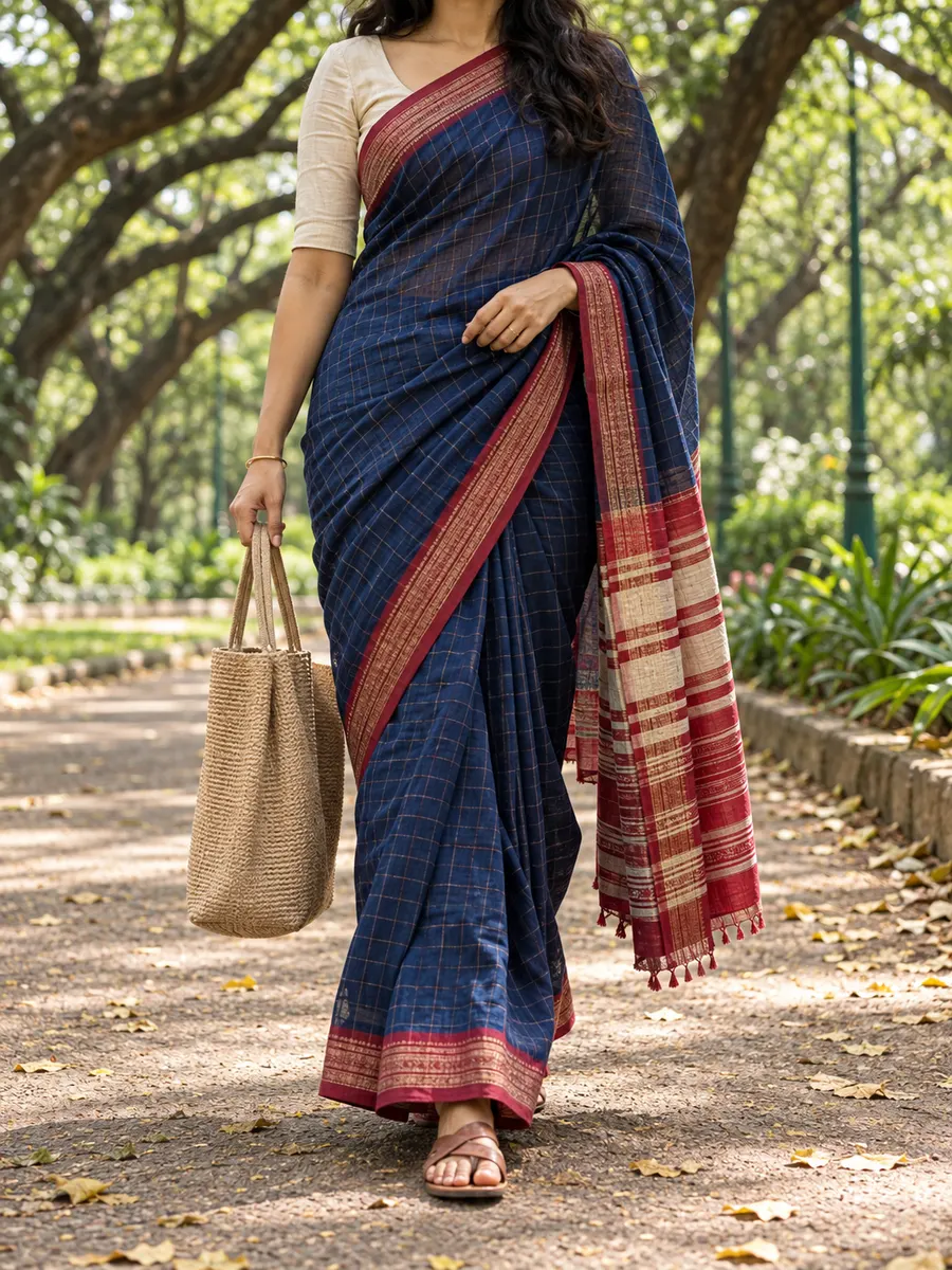A woman in an indigo Ilkal saree with a red-checked pallu, carrying a jute tote along a tree-lined path in a park.